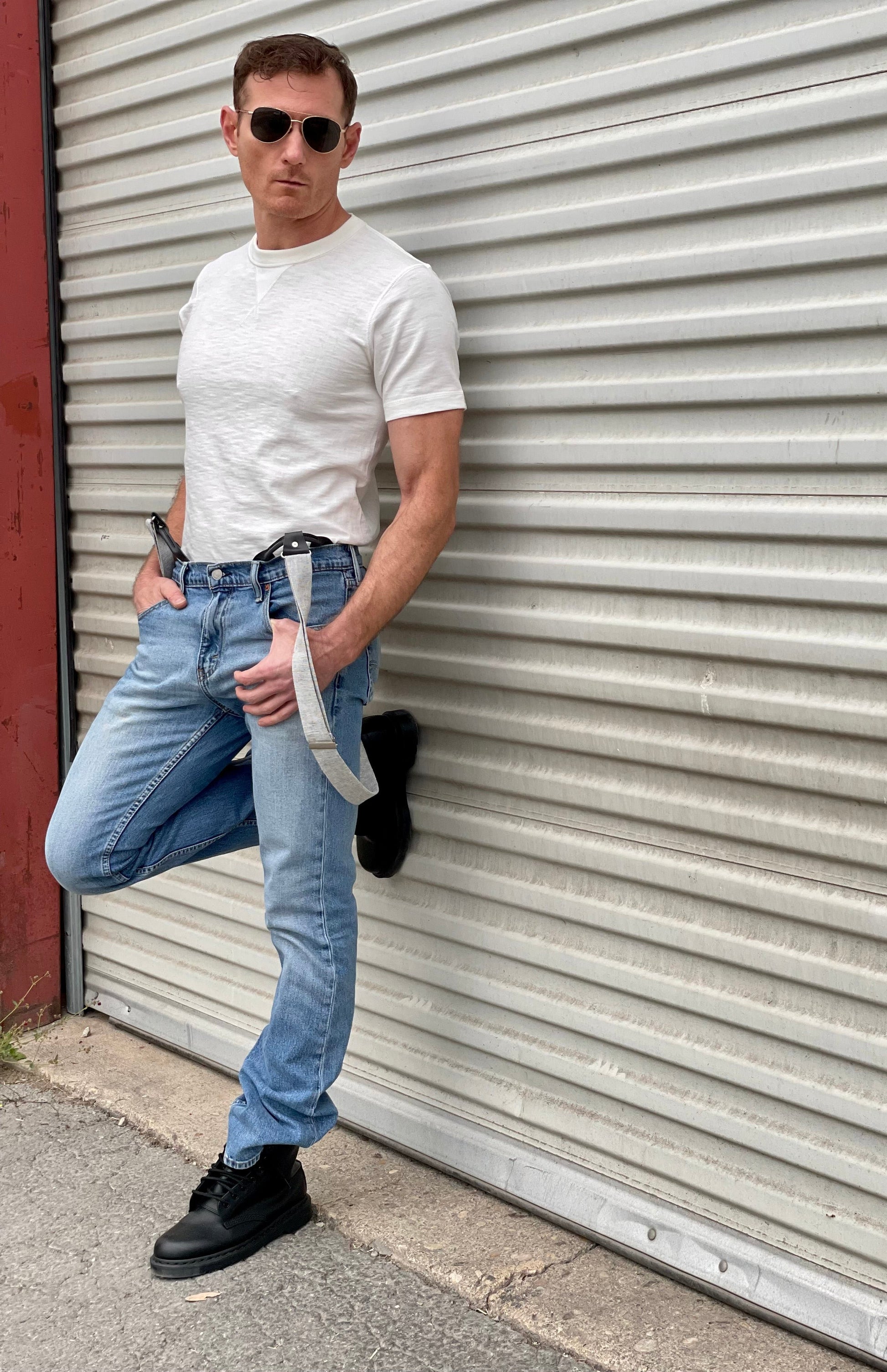 Man wearing a vintage-inspired 1940s heavyweight white t-shirt with gray linen suspenders by Stratton Suspender Co. Styled with classic denim and rugged black boots, this heritage menswear look is captured against an industrial metal backdrop, embodying timeless American workwear.