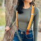 Close-up of women’s tangerine orange linen suspenders with tan leather magnetic clasps styled with dark denim jeans and fitted ribbed top, photographed outdoors by a tree near water.