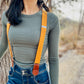 Close-up of women’s tangerine orange linen suspenders with tan leather magnetic clasps styled with dark denim jeans and fitted ribbed top in a rustic outdoor setting. 
