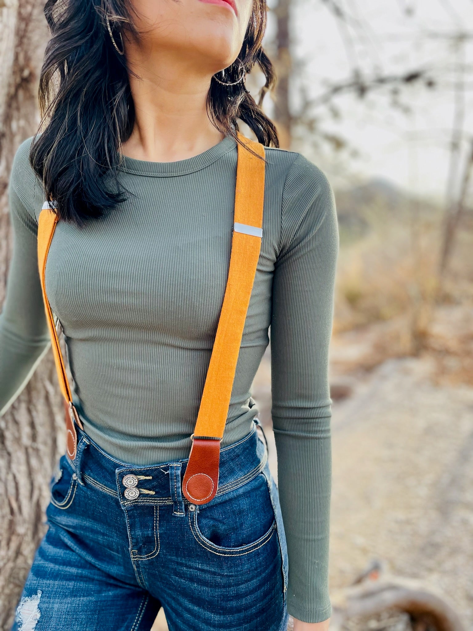 Close-up of women’s tangerine orange linen suspenders with tan leather magnetic clasps styled with dark denim jeans and fitted ribbed top in a rustic outdoor setting. 