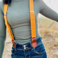 Close-up of women’s tangerine orange linen suspenders with tan leather magnetic clasps styled with high-waist jeans and ribbed top.