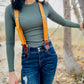 Women’s tangerine orange linen suspenders with tan magnetic clasps styled with distressed denim and ribbed top in a fall outdoor setting.