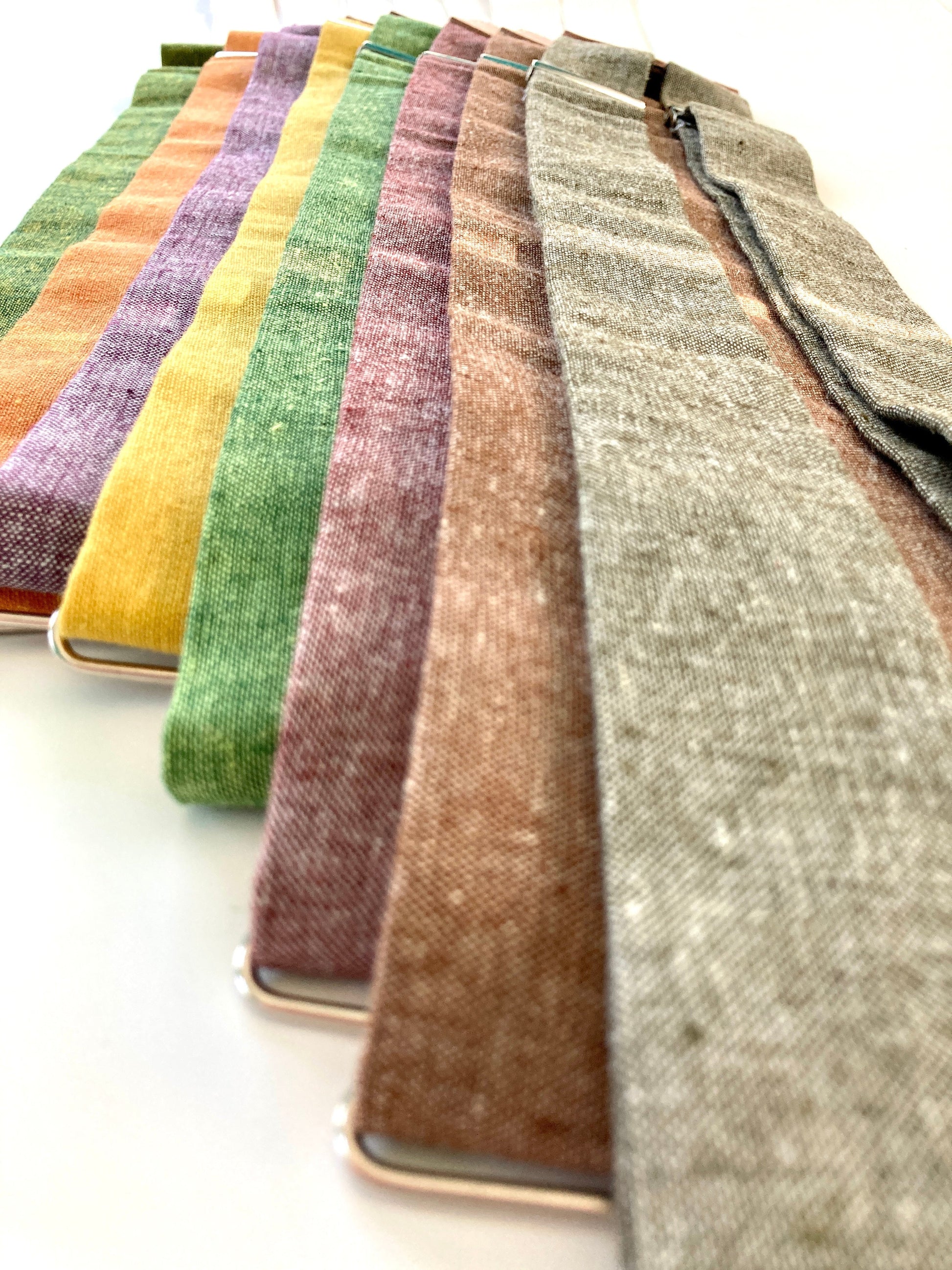 Stratton Suspender Co. Fall Linen Collection straps laid out in the following order: Palm (green and yellow woven linen), Orange, Purple, Yellow, Spruce (Bright Green), Rust or Maroon, Nutmeg (Brown), Olive Green
