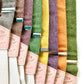 Stratton Suspender Co. Fall Linen Collection straps laid out in the following order: Spruce (bright green), Nutmeg (brown), Yellow, Maroon (rust), Orange, Olive Green, Palm Green (green and yellow woven linen), Purple 