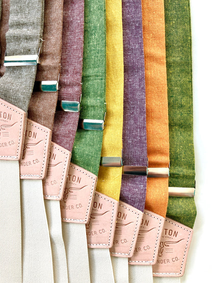 Stratton Suspender Co. Fall Linen Collection straps laid out in the following order: Spruce (bright green), Nutmeg (brown), Yellow, Maroon (rust), Orange, Olive Green, Palm Green (green and yellow woven linen), Purple 